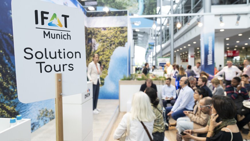 Hochgehaltenes Schild der IFAT Munich Solution Tours, dahinter sind unscharf einige Menschen zu sehen die einem Vortrag an einem Messestand lauschen.