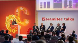 Fünf Männer in Anzügen sitzen auf einer Bühne der ceramitec talks und sprechen zu einem interessierten Publikum. Eine Frau moderiert den Talk.
