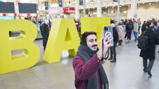 Ein lächelnder Mann in einer roter Jacke und grauem Schal steht vor den großen gelben Buchstaben „BAU“ und macht ein Selfie.