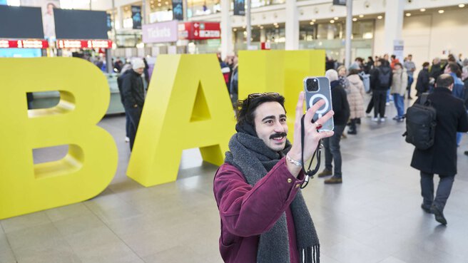 A smiling man in a red jacket and gray scarf stands in front of the large yellow letters 