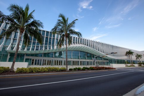 The exterior façade of the MIAMI BEACH CONVENTION CENTRE with numerous entrance doors, a curved canopy, palm trees, an empty street and a slightly cloudy sky.