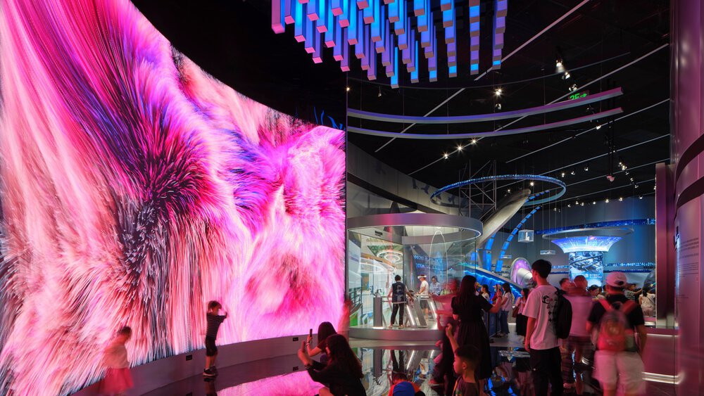 A futuristic exhibition area with a huge screen, columns, arches and information stands, bustling with visitors.