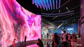 A futuristic exhibition area with a huge screen, columns, arches and information stands, bustling with visitors.