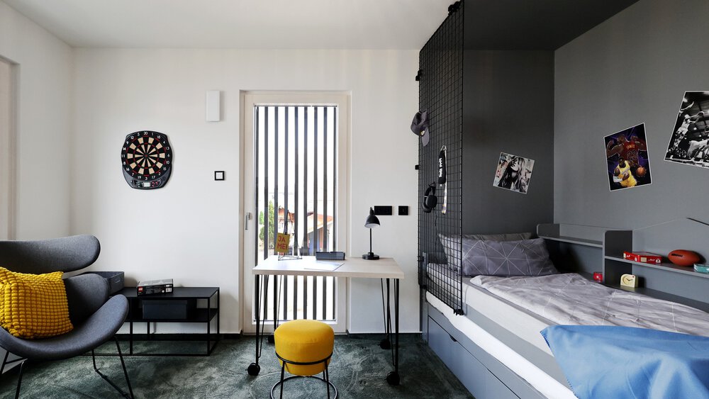 Ein modernes Jugendzimmer mit einem Bett, Tisch, Stühlen, einem Regal, einer Dartscheibe und einer Balkontür.