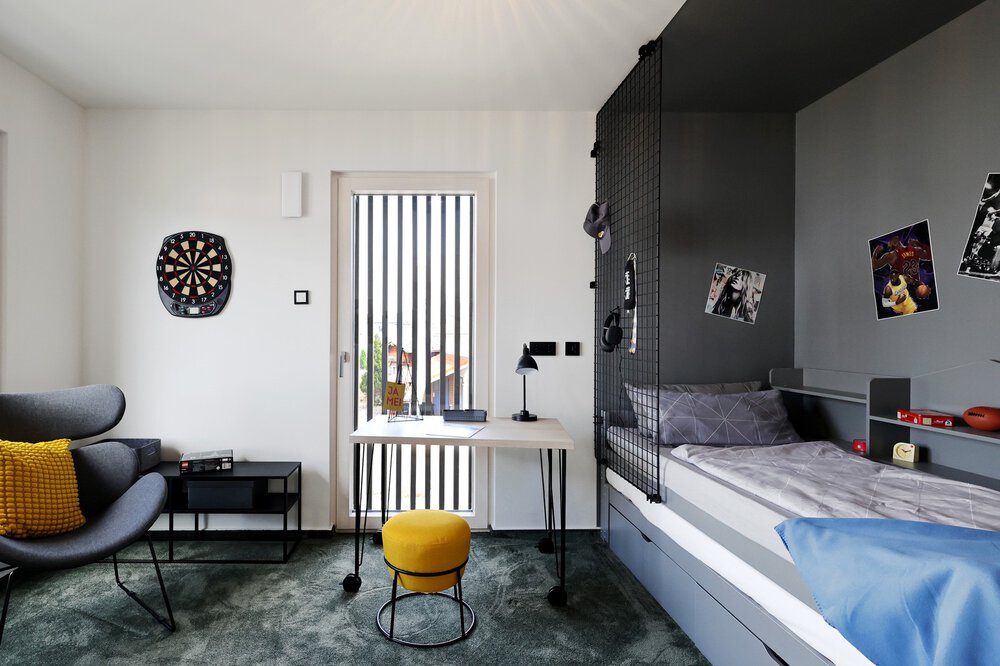 Ein modernes Jugendzimmer mit einem Bett, Tisch, Stühlen, einem Regal, einer Dartscheibe und einer Balkontür.