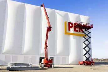 Two aerial work platforms are being used on the modern white building facade of the PERI building at bauma. A scissor lift is used to attach letters, while a telescopic boom lift reaches the top edge of the building. Construction materials are in the foreground, and the sky is clear and blue.