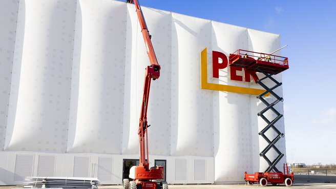 Zwei Hebebühnen werden an der modernen weißen Gebäudefassade des PERI-Gebäudes auf der bauma eingesetzt. Mithilfe einer Scherenbühne werden Buchstaben befestigt, während eine Teleskopbühne bis zum oberen Rand des Gebäudes reicht. Im Vordergrund liegen Baumaterialien, und der Himmel ist klar und blau.