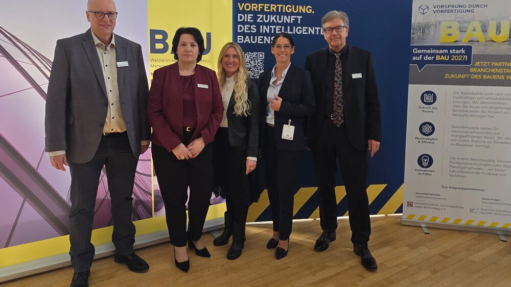 Fünf Personen in Business-Kleidung stehen lächelnd vor Bannern auf einer professionellen Veranstaltung.