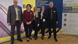 Fünf Personen in Business-Kleidung stehen lächelnd vor Bannern auf einer professionellen Veranstaltung.