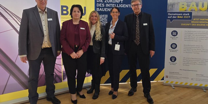 Fünf Personen in Business-Kleidung stehen lächelnd vor Bannern auf einer professionellen Veranstaltung.