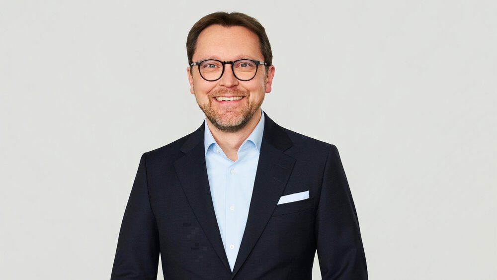 A smiling man with short brown hair, glasses and a beard, wearing a light blue shirt with a black jacket and white pocket square.
