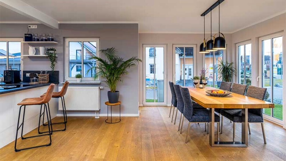 Moderner Essbereich mit Holztisch, grauen Stühlen, Barhockern an einer Theke und großen Fenstern mit Blick auf eine Terrasse.