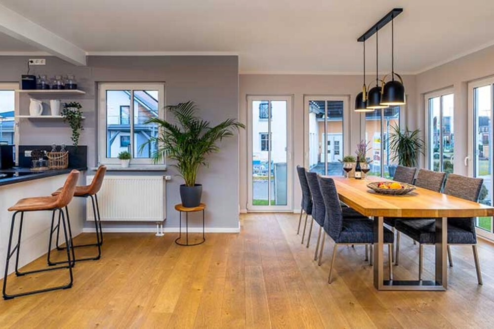 Moderner Essbereich mit Holztisch, grauen Stühlen, Barhockern an einer Theke und großen Fenstern mit Blick auf eine Terrasse.