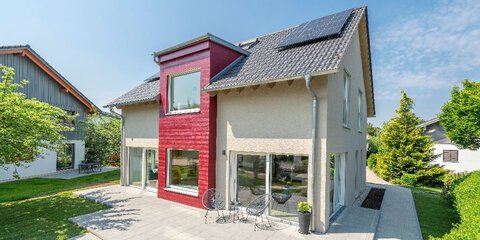 Ein Musterhaus mit steilem Dach, beiger und roter Hauswand, bodentiefen Fenstern und einer großen Terrasse mit Sitzmöbeln.