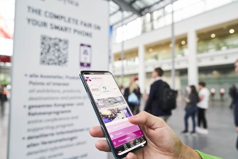 A hand holding a smartphone with an events app open, in a bright, modern building with people in the background.