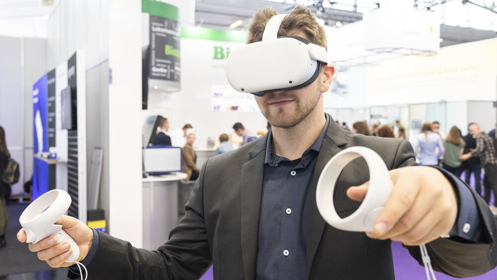A man in a suit wears white VR goggles and holds two white round controllers in his hands. An exhibition stand can be seen in the background