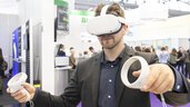A man in a suit wears white VR goggles and holds two white round controllers in his hands. An exhibition stand can be seen in the background