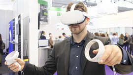 A man in a suit wears white VR goggles and holds two white round controllers in his hands. An exhibition stand can be seen in the background