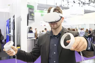 A man in a suit wears white VR goggles and holds two white round controllers in his hands. An exhibition stand can be seen in the background