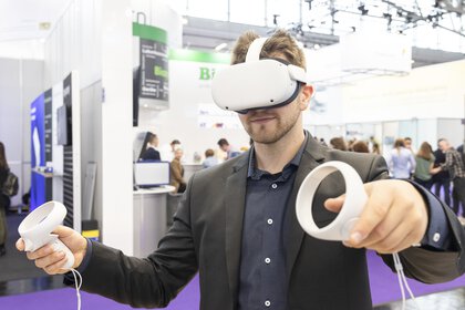 A man in a suit wears white VR goggles and holds two white round controllers in his hands. An exhibition stand can be seen in the background