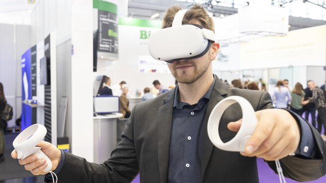 A man in a suit wears white VR goggles and holds two white round controllers in his hands. An exhibition stand can be seen in the background