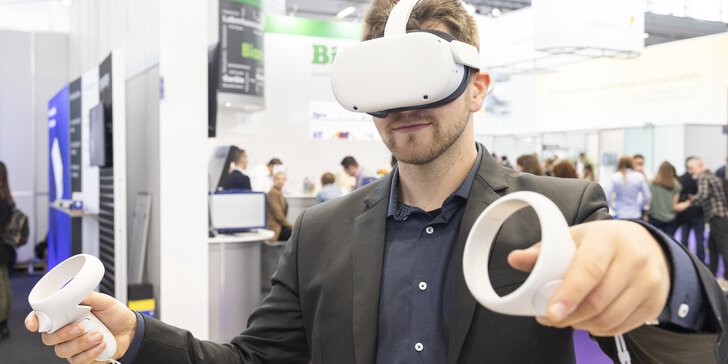 A man in a suit wears white VR goggles and holds two white round controllers in his hands. An exhibition stand can be seen in the background