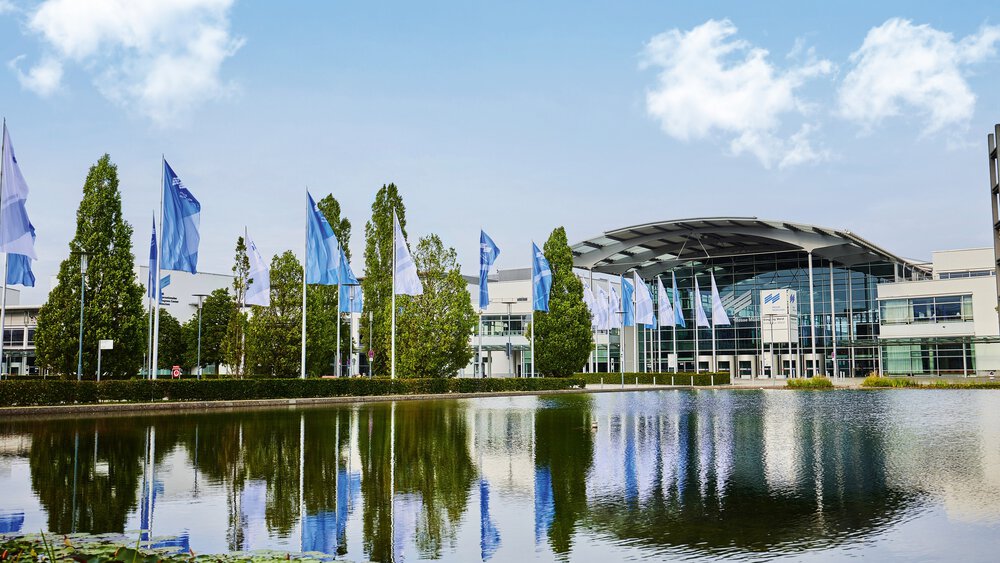 Der Eingang West des Messe München Geländes mit dem Messesee im Vordergrund und einer Allee aus Fahnen und Bäumen, die auf die Eingangshalle zuführt.