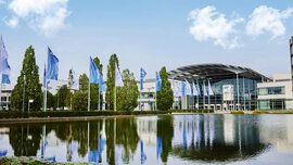 Der Eingang West des Messe München Geländes mit dem Messesee im Vordergrund und einer Allee aus Fahnen und Bäumen, die auf die Eingangshalle zuführt.