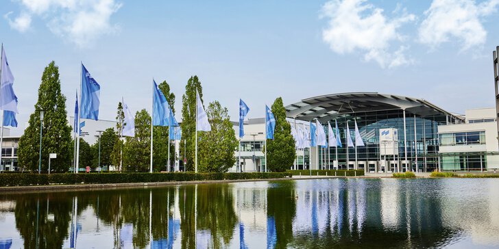 Der Eingang West des Messe München Geländes mit dem Messesee im Vordergrund und einer Allee aus Fahnen und Bäumen, die auf die Eingangshalle zuführt.
