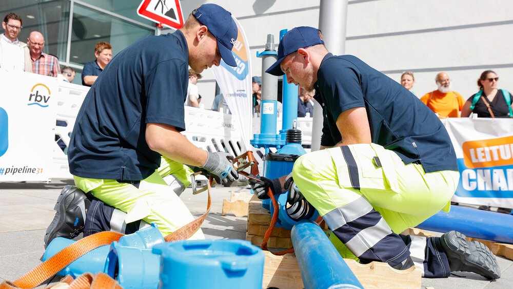 Zwei Personen befestigen ein blaues Rohrleitungsbauteil mit Spanngurten auf Paletten bei der Leitungsbau Challenge, im Hintergrund Absperrungen und Banner.