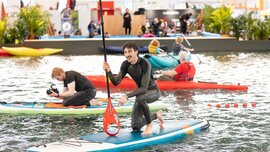 A man is kneeling on a stand-up paddleboard in the swimming pool of an trade fair hall.
