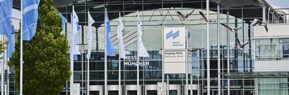 A modern exhibition centre with a glass façade, blue flags and birds in flight, in front of the Munich Trade Fair Centre, Germany.