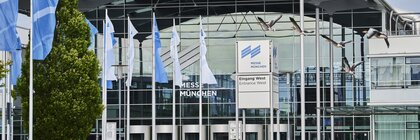 Modernes Messegelände mit Glasfassade, blauen Fahnen und fliegenden Vögeln vor der Messe München, Deutschland