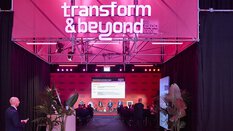 A panel discussion takes place on the “Transform & Beyond by EXPO REAL” stage in Munich. Several speakers are seated on stage in front of an audience, with a large screen behind them displaying the event program. The stage is illuminated in pink lighting, surrounded by black curtains and plants, creating a modern and dynamic atmosphere.