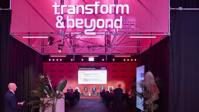 Eine Podiumsdiskussion auf der Bühne „Transform & Beyond by EXPO REAL“ in München. Mehrere Sprecherinnen und Sprecher sitzen auf der Bühne vor einem Publikum, während hinter ihnen eine große Leinwand mit dem Veranstaltungsprogramm angezeigt wird. Die Bühne ist in pinkem Licht beleuchtet, umgeben von schwarzen Vorhängen und Pflanzen, und vermittelt eine moderne, dynamische Atmosphäre.