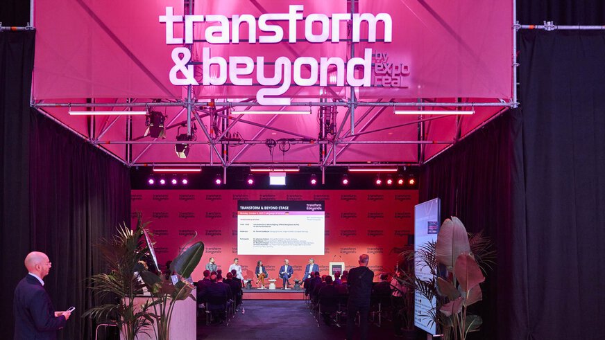 A panel discussion takes place on the “Transform & Beyond by EXPO REAL” stage in Munich. Several speakers are seated on stage in front of an audience, with a large screen behind them displaying the event program. The stage is illuminated in pink lighting, surrounded by black curtains and plants, creating a modern and dynamic atmosphere.