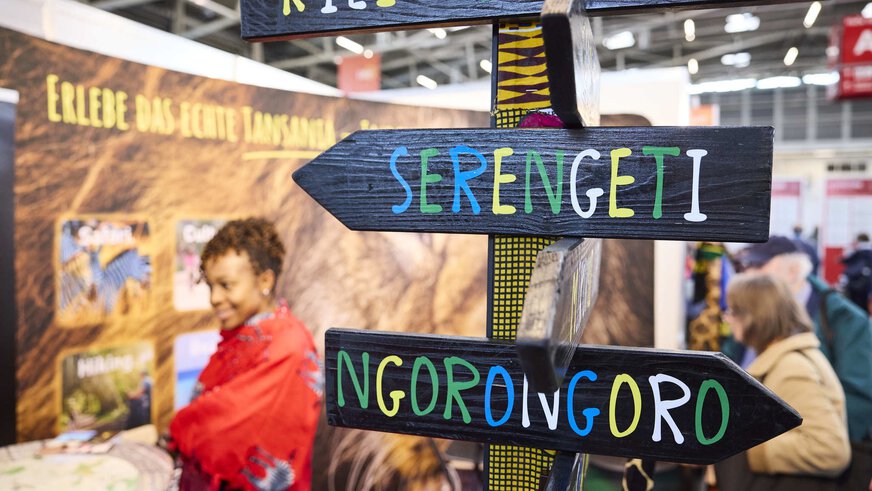 Wooden signposts with “Kilimanjaro”, “Serengeti” and “Ngorongoro” at the f.re.e trade fair, in front of a Tanzania stand with a smiling woman.