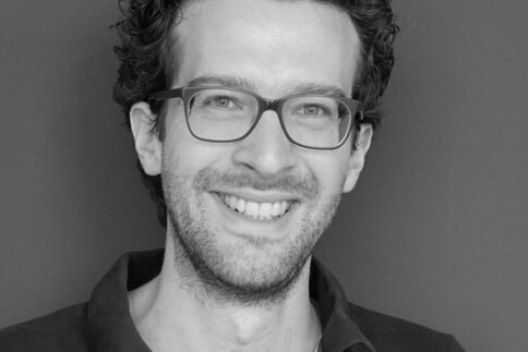 Schwarzweißfoto eines lächelnden jungen Mannes mit kurzem, dunklem welligen Haar, einer Brille und Bart.