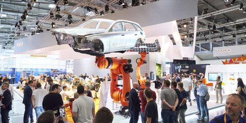 Visitors observe a large industrial robot lifting a silver car body at a trade fair.