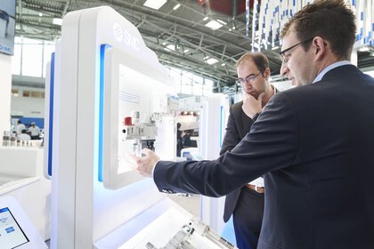 Zwei Männer in Anzügen untersuchen ein High-Tech-Ausstellungsstück an einem Indoor-Messestand.