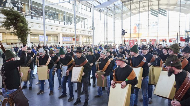 Blick in die menschengefüllte Eingangshalle eines Messegebäudes. Vorne treten Personen in Tracht mit einer traditionellen Vorführung mit großen Glocken auf.