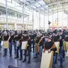 View into the crowded entrance hall of a trade fair building. People with large bells appear in front.