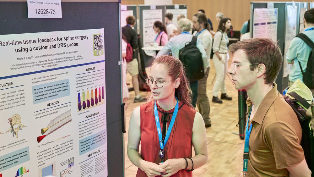 Two people stand together in front of an information wall and look at a poster. In the background, you can see other walls and visitors.