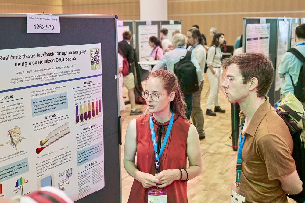 Two people stand together in front of an information wall and look at a poster. In the background, you can see other walls and visitors.