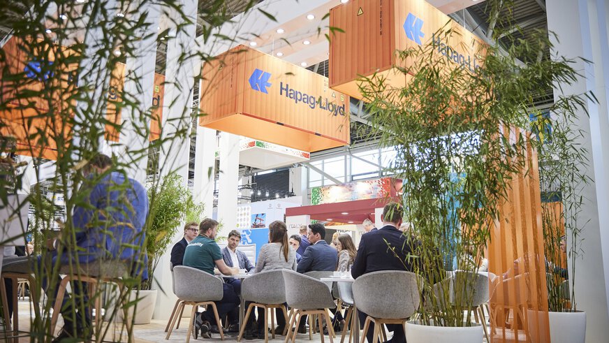 Eine Gruppe Menschen sitzt um einen Tisch, rechts und links davon stehen Pflanzen und an der Hallendecke hängen Werbebanner in der Form von orangen Frachtcontainern.