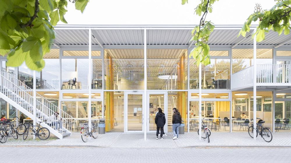 Modernes zweistöckiges Gebäude mit Glasfassade, draußen geparkten Fahrrädern und Menschen, die in der Nähe unter grünen Blättern spazieren gehen.