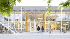 Modernes zweistöckiges Gebäude mit Glasfassade, draußen geparkten Fahrrädern und Menschen, die in der Nähe unter grünen Blättern spazieren gehen.