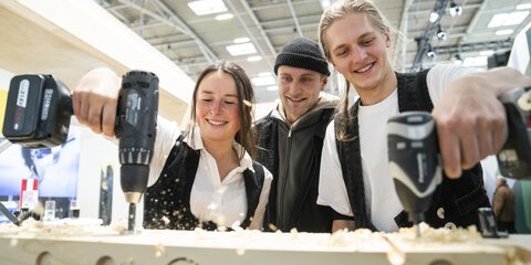 Drei lächelnde Personen stehen vor einem Holzblock in einer Halle der BAU. Zwei der Personen halten jeweils eine Bohrmaschine in der Hand und bohren ins Holz.