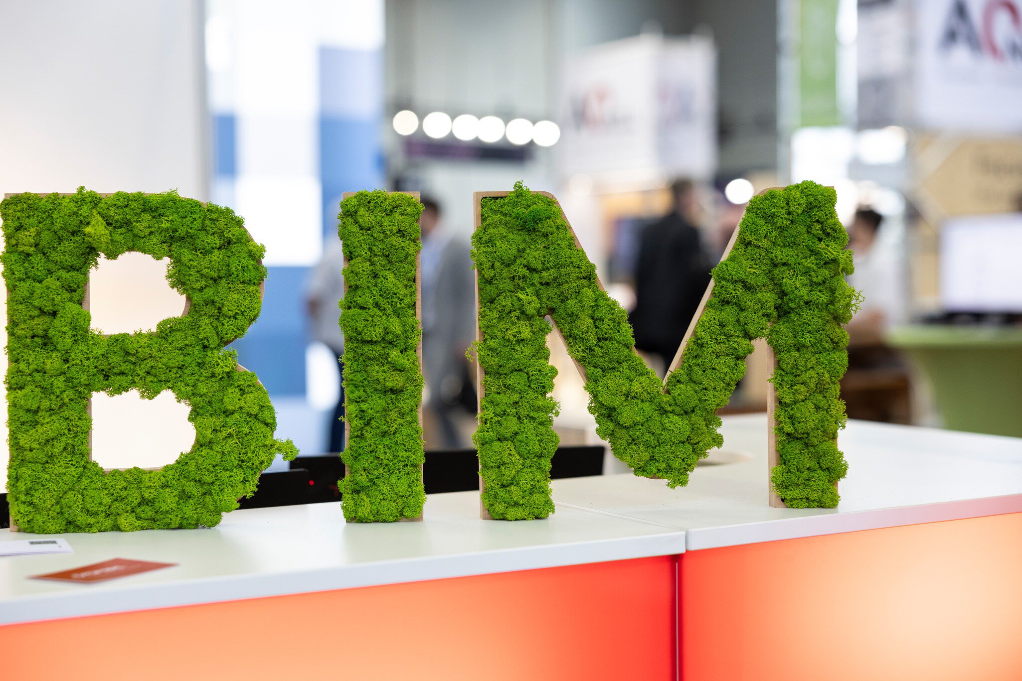 Der Schriftzug „BIM“ in hölzernen Buchstaben, aus denen vorne grüne Pflanzen herauswachsen. Im Hintergrund sind unscharf Messestände zu erkennen.