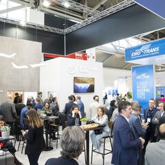 Visitors to transport logistic sit and stand at tables at a trade fair stand. Other exhibition stands can be seen in the background.